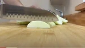 Close-up of a knife slicing green apple wedges on a wooden cutting board in a kitchen setting.
